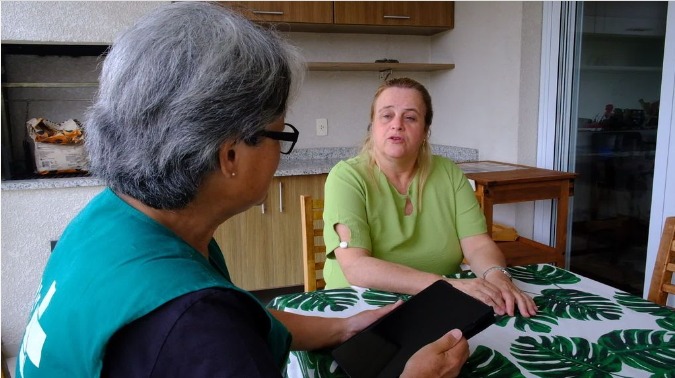A foto mostra duas mulheres sentadas à mesa de uma cozinha; uma delas está de perfil, veste um colete verde, possui cabelo grisalho e segura um tablet nas mãos; a outra aparece de frente, possui cabelo louto, veste uma blusa verde e está falando